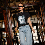 A woman walking through a grand, dark wood doorway wearing a black graphic t-shirt, light-wash blue jeans, sunglasses, and carrying a small gold handbag.