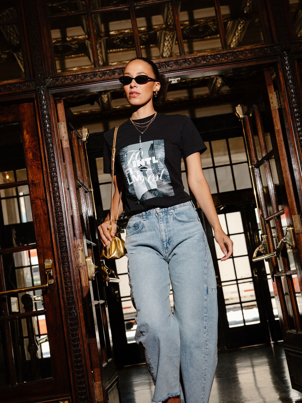A woman walking through a grand, dark wood doorway wearing a black graphic t-shirt, light-wash blue jeans, sunglasses, and carrying a small gold handbag.