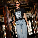 A woman walking through a grand, dark wood doorway wearing a black graphic t-shirt, light-wash blue jeans, sunglasses, and carrying a small gold handbag.