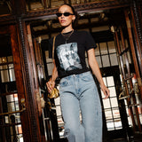 A woman walking through a grand, dark wood doorway wearing a black graphic t-shirt, light-wash blue jeans, sunglasses, and carrying a small gold handbag.