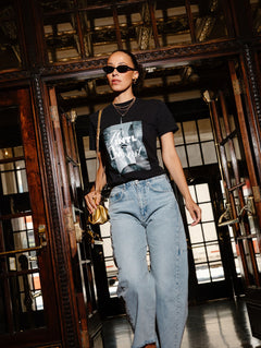 A woman walking through a grand, dark wood doorway wearing a black graphic t-shirt, light-wash blue jeans, sunglasses, and carrying a small gold handbag.