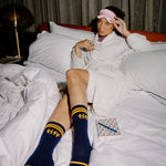 A woman reclines on a bed wearing a white robe, pink sleep mask, and navy blue socks with yellow stripes and the text 'tiff' on them. A small patterned notebook lies beside her on the white bedding.