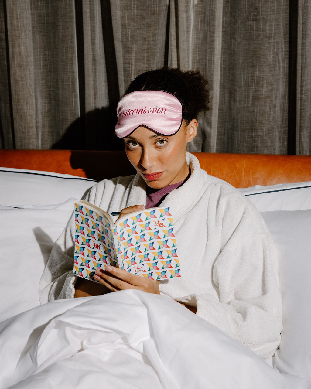 A woman in a white robe sits in bed under white covers, wearing a pink sleep mask with the word 'intermission' embroidered on it and holding an open book with a colorful geometric pattern.