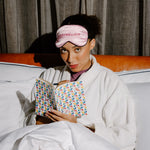 A woman in a white robe sits in bed under white covers, wearing a pink sleep mask with the word 'intermission' embroidered on it and holding an open book with a colorful geometric pattern.