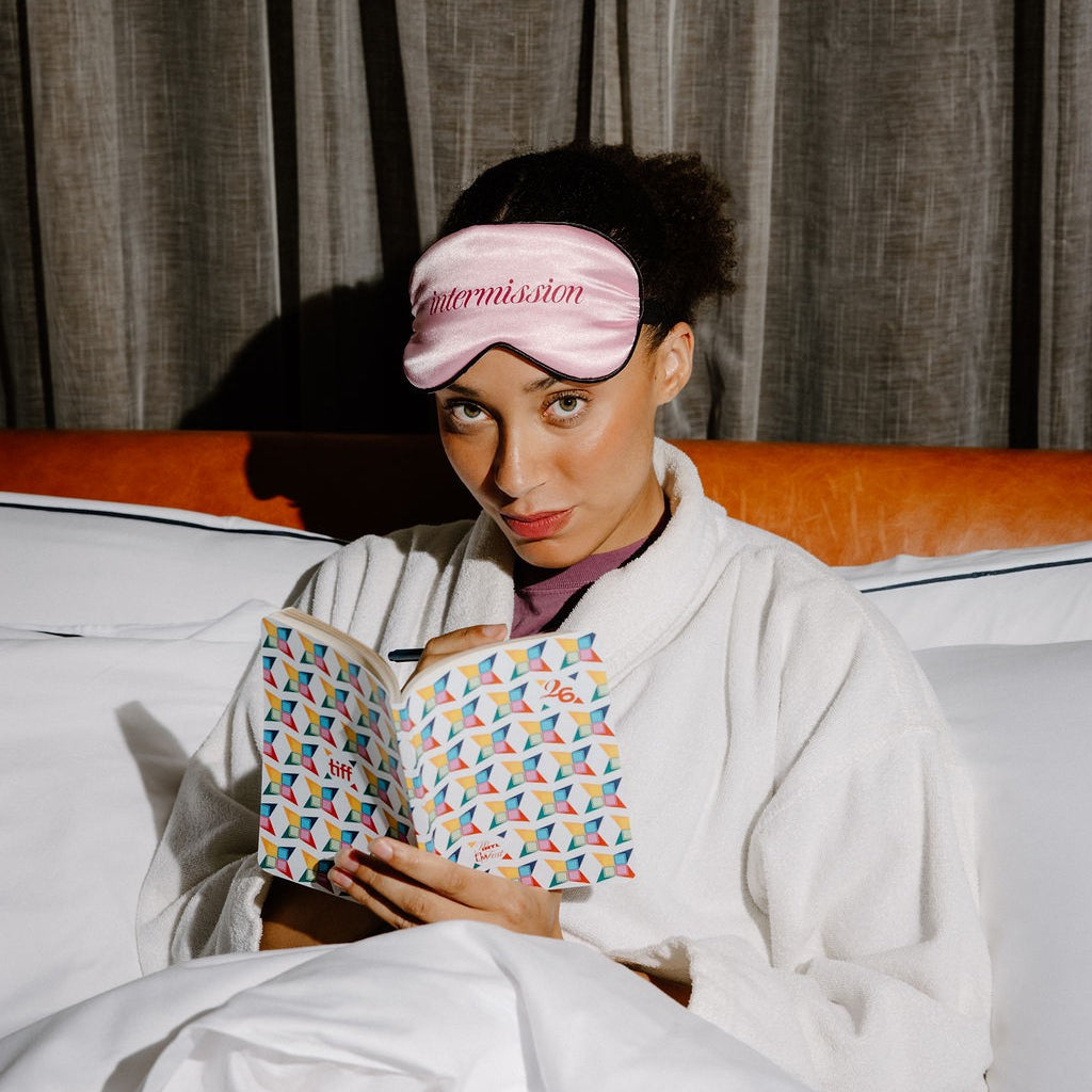 A woman in a white robe sits in bed under white covers, wearing a pink sleep mask with the word 'intermission' embroidered on it and holding an open book with a colorful geometric pattern.