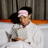 A woman in a white robe sits in bed under white covers, wearing a pink sleep mask with the word 'intermission' embroidered on it and holding an open book with a colorful geometric pattern.