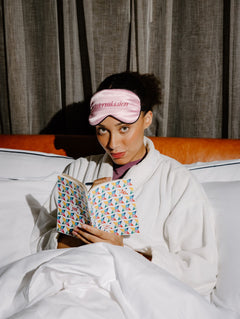 A woman in a white robe sits in bed under white covers, wearing a pink sleep mask with the word 'intermission' embroidered on it and holding an open book with a colorful geometric pattern.