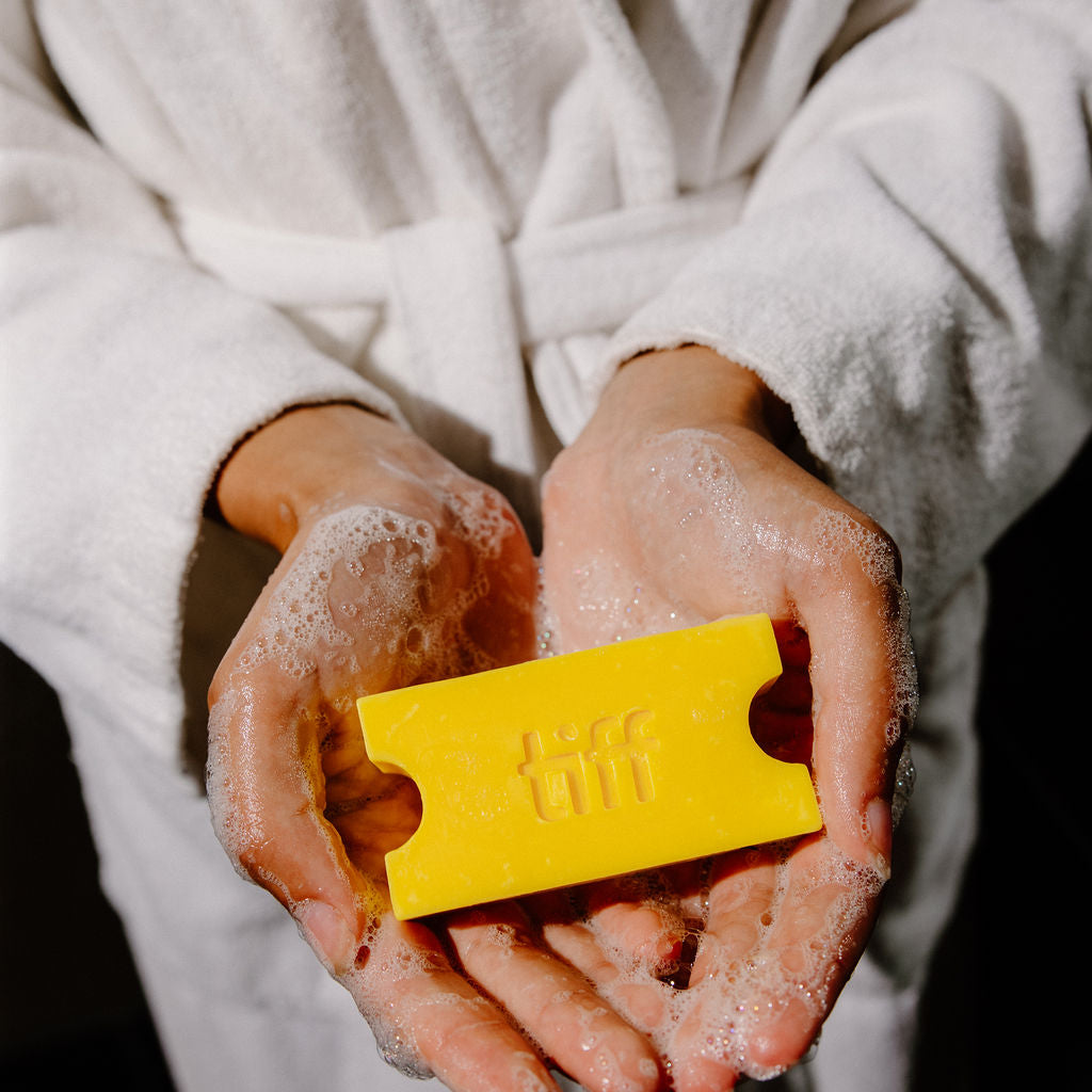 A person in a white bathrobe holds a bright yellow rectangular soap bar with embossed lettering in their hands, which are covered in white foam, against a dark background.