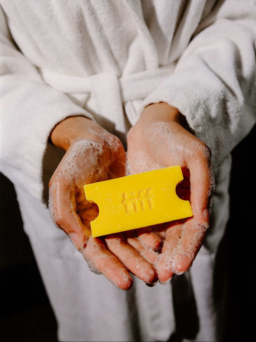 A person in a white bathrobe holds a bright yellow rectangular soap bar with embossed lettering in their hands, which are covered in white foam, against a dark background.
