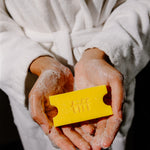 A person in a white bathrobe holds a bright yellow rectangular soap bar with embossed lettering in their hands, which are covered in white foam, against a dark background.