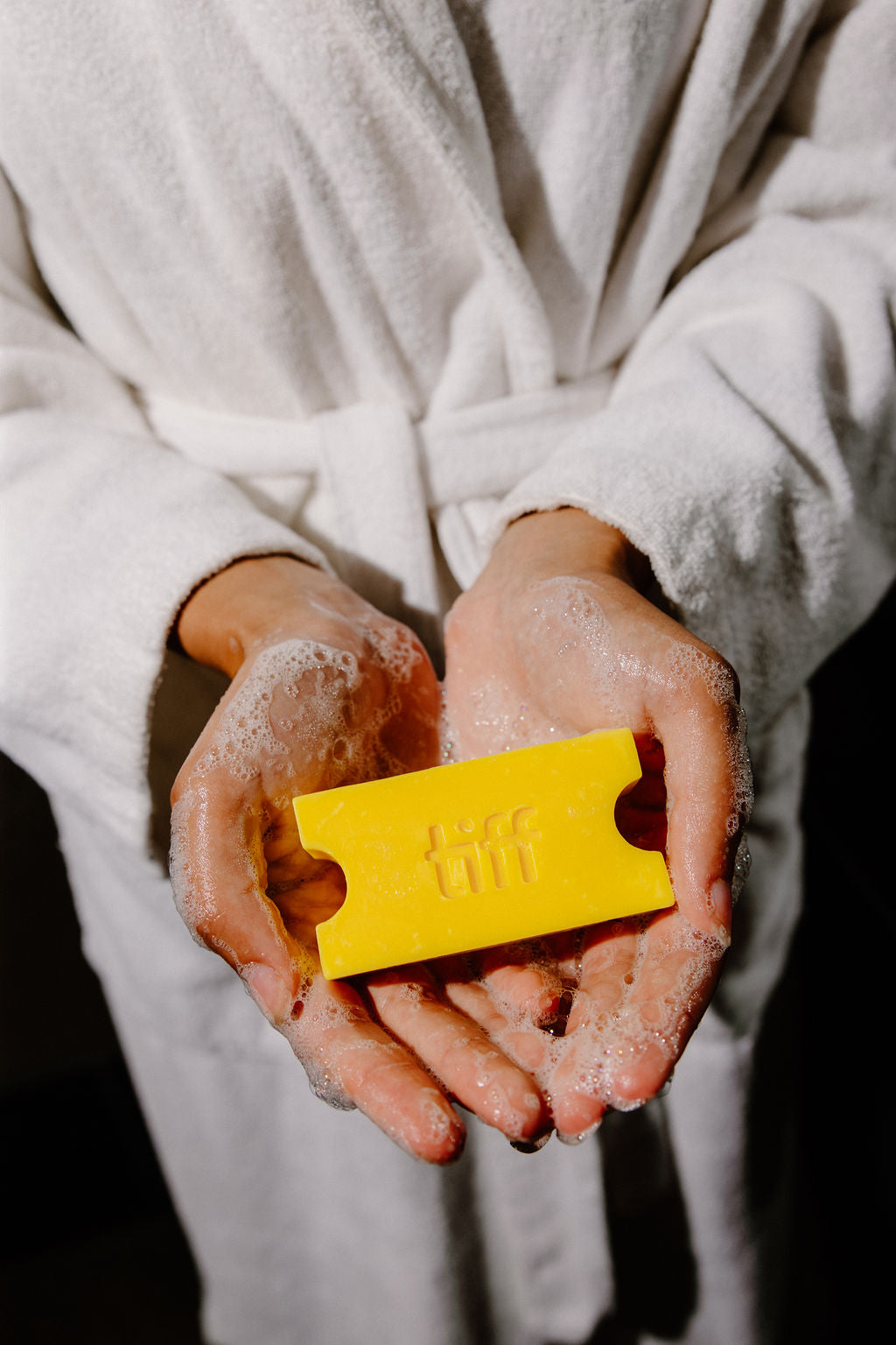 A person in a white bathrobe holds a bright yellow rectangular soap bar with embossed lettering in their hands, which are covered in white foam, against a dark background.