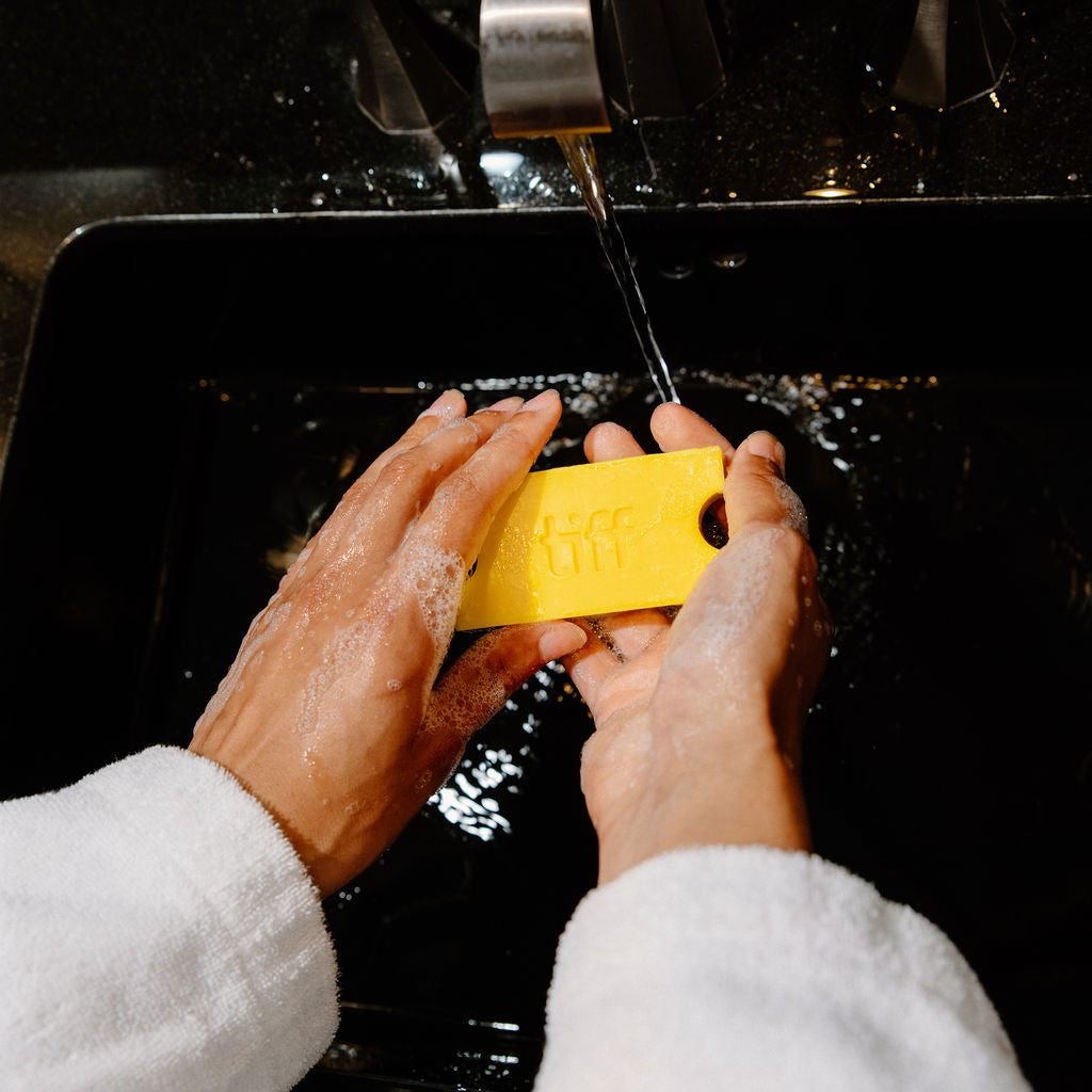 A person wearing a white bathrobe washes a yellow rectangular bar of soap under running water in a black sink.