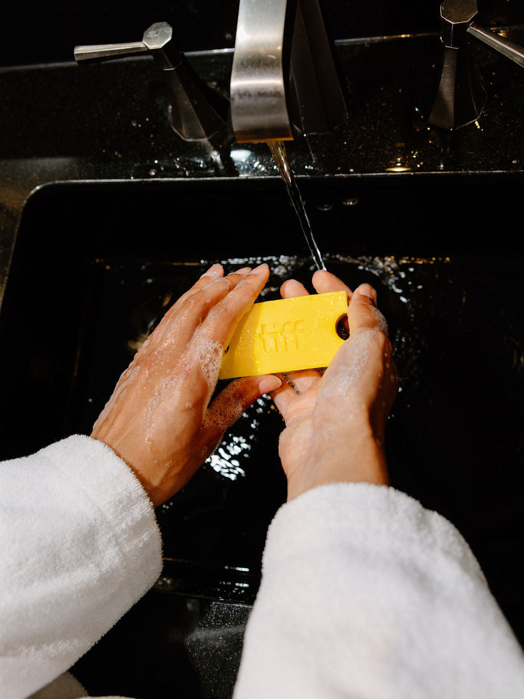 A person wearing a white bathrobe washes a yellow rectangular bar of soap under running water in a black sink.