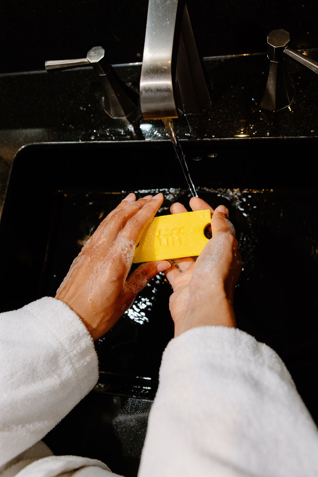 A person wearing a white bathrobe washes a yellow rectangular bar of soap under running water in a black sink.