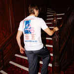 A man wearing a white graphic t-shirt and dark trousers walks up a wooden staircase with a red patterned carpet, viewed from behind to show the back of the t-shirt.