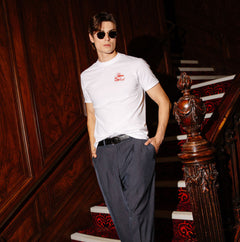 A man wearing a white short-sleeve t-shirt with a small red logo on the left chest, dark gray trousers, and sunglasses, posing on an ornate wooden staircase with a red patterned carpet.
