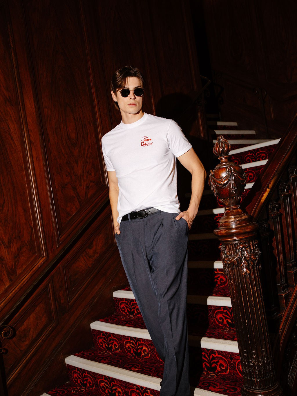 A man wearing a white short-sleeve t-shirt with a small red logo on the left chest, dark gray trousers, and sunglasses, posing on an ornate wooden staircase with a red patterned carpet.
