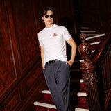 A man wearing a white short-sleeve t-shirt with a small red logo on the left chest, dark gray trousers, and sunglasses, posing on an ornate wooden staircase with a red patterned carpet.