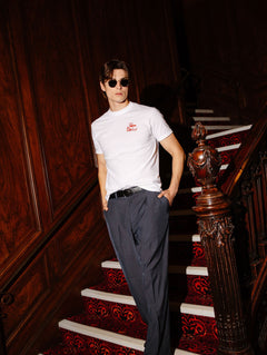 A man wearing a white short-sleeve t-shirt with a small red logo on the left chest, dark gray trousers, and sunglasses, posing on an ornate wooden staircase with a red patterned carpet.