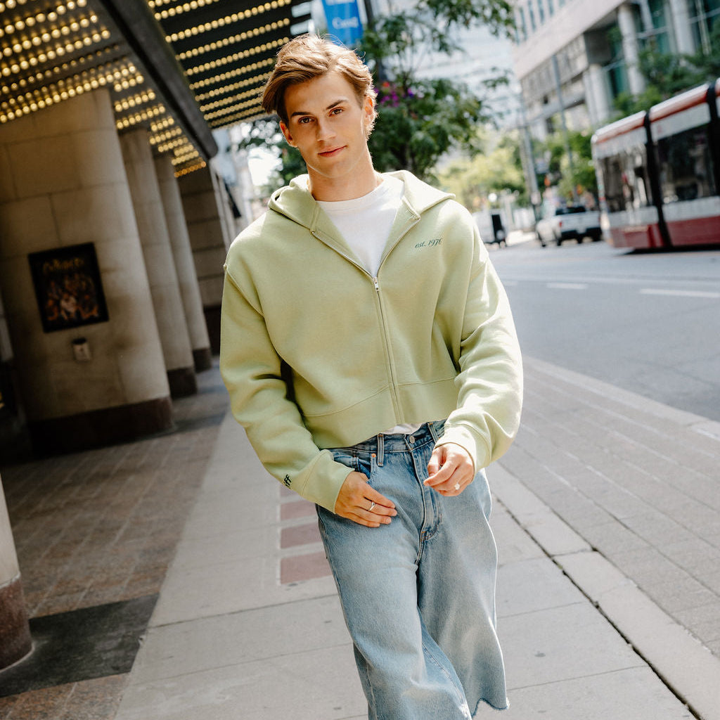 A young man walking on a city sidewalk wearing a light sage green zip-up hoodie, light-wash denim shorts, and green and white sneakers.
