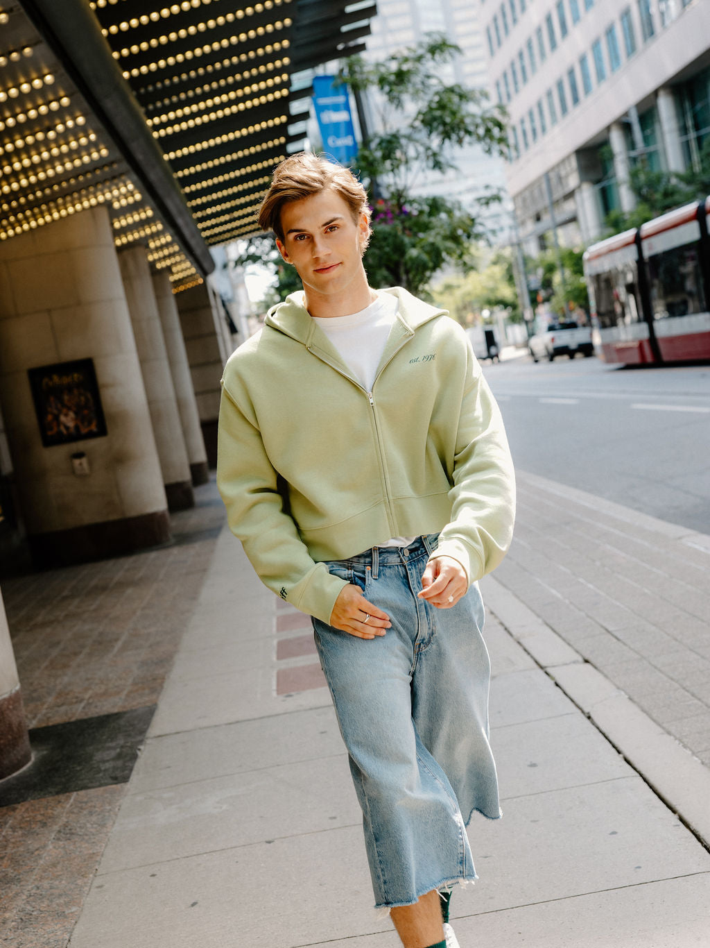 A young man walking on a city sidewalk wearing a light sage green zip-up hoodie, light-wash denim shorts, and green and white sneakers.