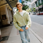 A young man walking on a city sidewalk wearing a light sage green zip-up hoodie, light-wash denim shorts, and green and white sneakers.