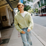 A young man walking on a city sidewalk wearing a light sage green zip-up hoodie, light-wash denim shorts, and green and white sneakers.