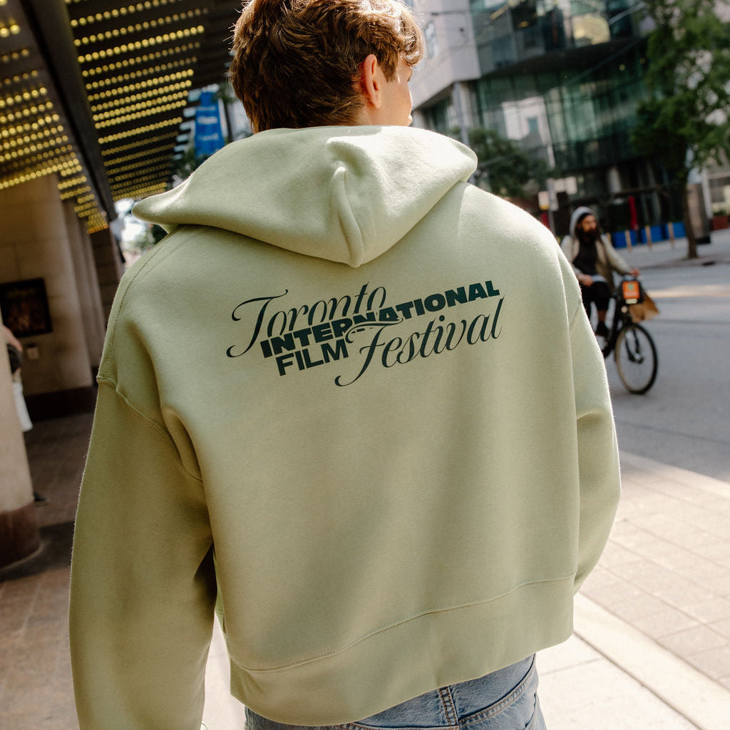 A person viewed from behind wearing a light sage green cropped hoodie with 'Toronto International Film Festival' printed in black across the back, paired with light blue jeans, standing on a city sidewalk.