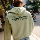 A person viewed from behind wearing a light sage green cropped hoodie with 'Toronto International Film Festival' printed in black across the back, paired with light blue jeans, standing on a city sidewalk.