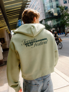 A person viewed from behind wearing a light sage green cropped hoodie with 'Toronto International Film Festival' printed in black across the back, paired with light blue jeans, standing on a city sidewalk.