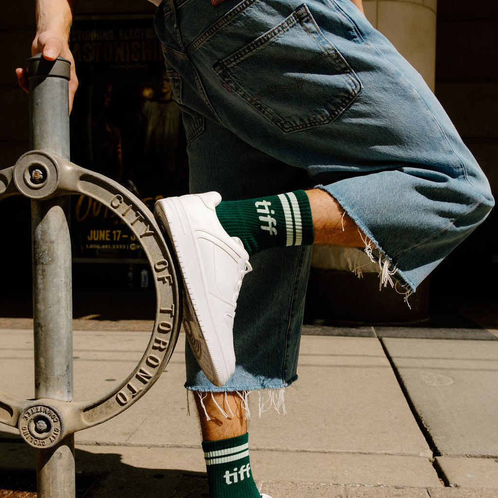 A person outdoors wearing light blue denim shorts with raw hems, white sneakers, and dark green crew socks with white stripes and the word 'tiff' printed on them.