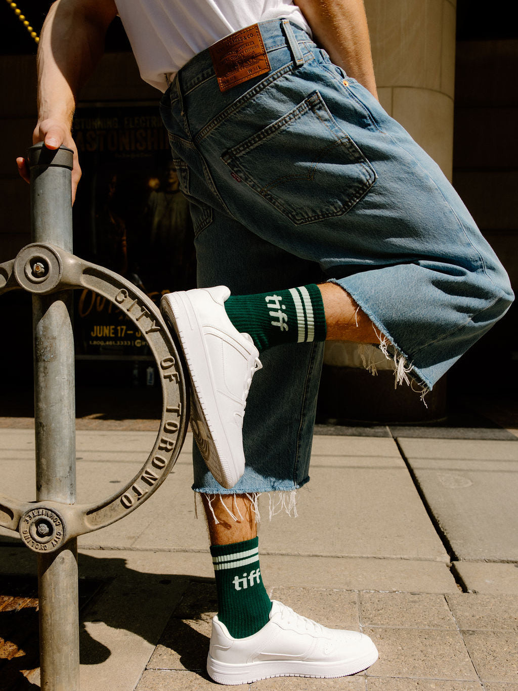 A person outdoors wearing light blue denim shorts with raw hems, white sneakers, and dark green crew socks with white stripes and the word 'tiff' printed on them.