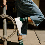 A person outdoors wearing light blue denim shorts with raw hems, white sneakers, and dark green crew socks with white stripes and the word 'tiff' printed on them.