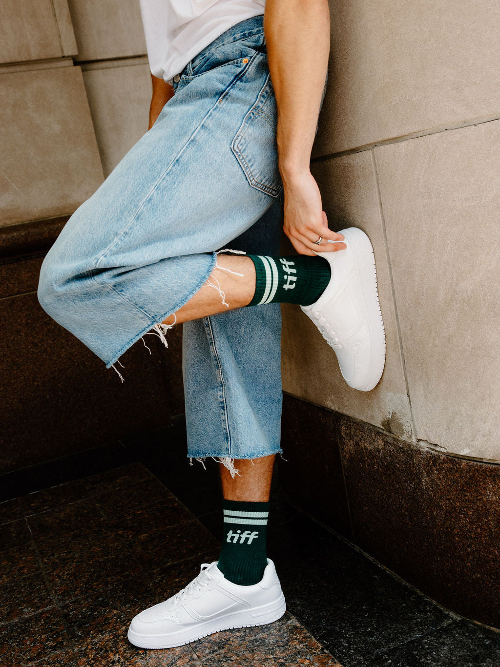 A person wearing light-wash frayed denim jeans, white sneakers, and dark green crew socks with white stripes and the word 'tiff' printed on them, leaning against a stone wall.