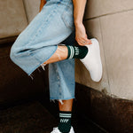 A person wearing light-wash frayed denim jeans, white sneakers, and dark green crew socks with white stripes and the word 'tiff' printed on them, leaning against a stone wall.