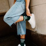 A person wearing light-wash frayed denim jeans, white sneakers, and dark green crew socks with white stripes and the word 'tiff' printed on them, leaning against a stone wall.