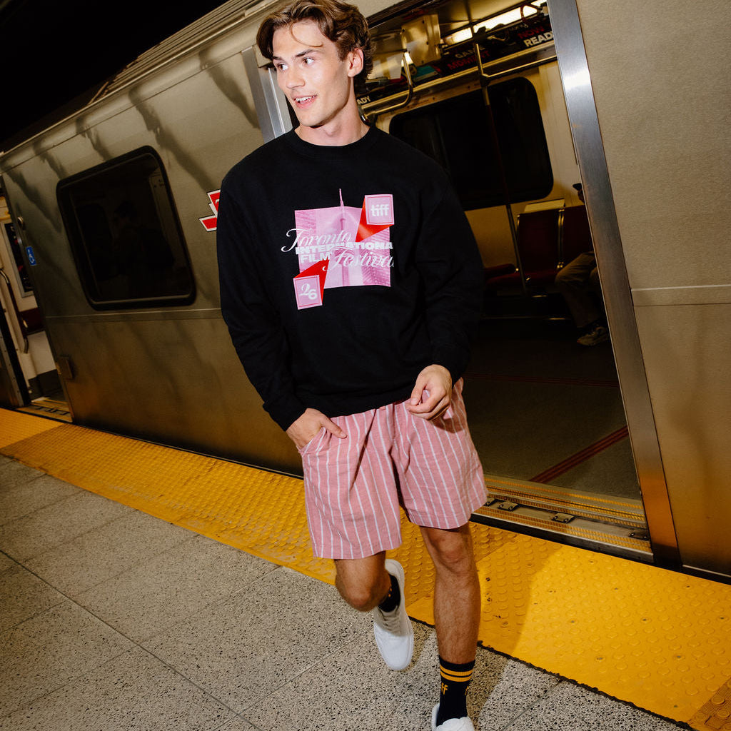A man walking off a train at a station, wearing a black crewneck sweatshirt with a pink and white graphic print, pink and white striped shorts, black and yellow socks, and white sneakers.