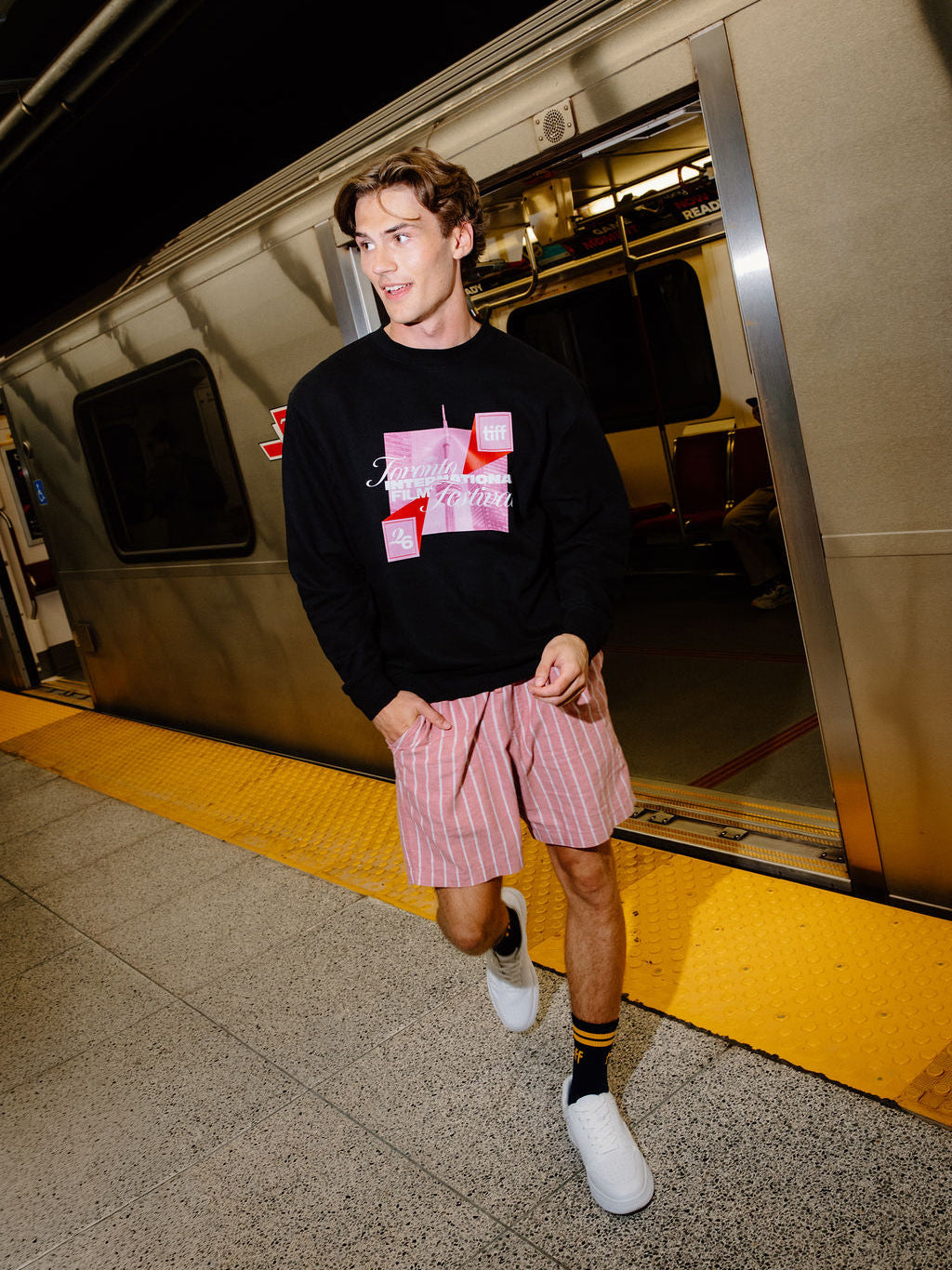 A man walking off a train at a station, wearing a black crewneck sweatshirt with a pink and white graphic print, pink and white striped shorts, black and yellow socks, and white sneakers.