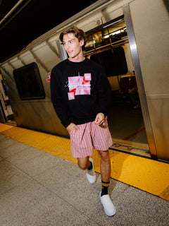 A man walking off a train at a station, wearing a black crewneck sweatshirt with a pink and white graphic print, pink and white striped shorts, black and yellow socks, and white sneakers.