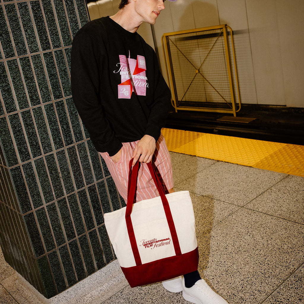 A person standing in a subway station holding a white canvas tote bag with a dark red bottom panel and handles. The person is wearing a black graphic sweatshirt, pink striped pants, white sneakers, and a purple cap.
