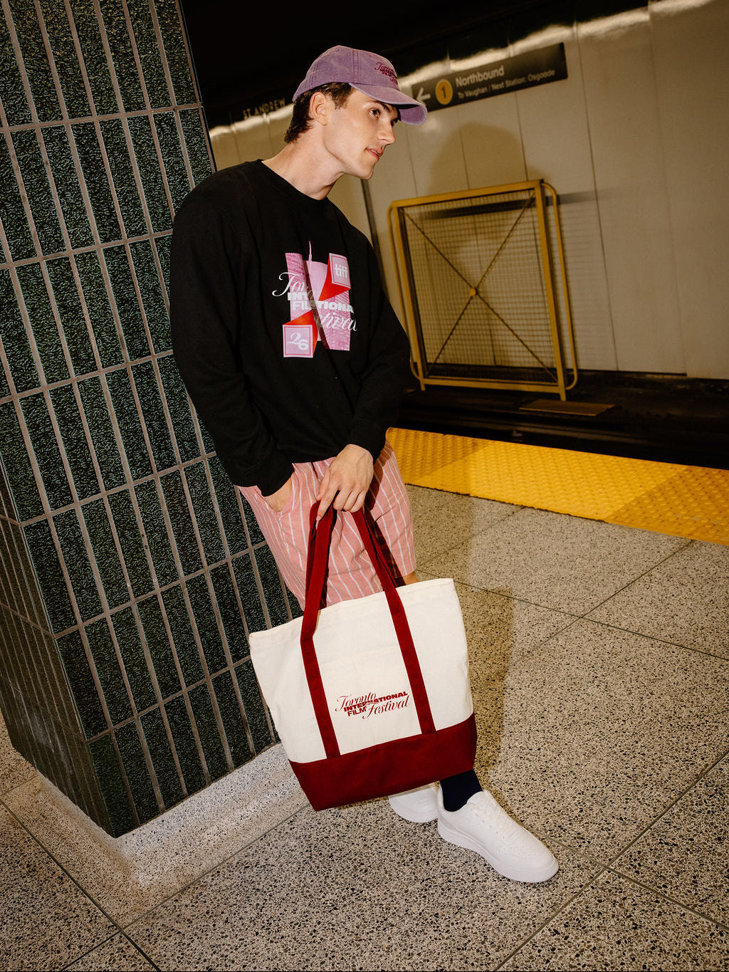A person standing in a subway station holding a white canvas tote bag with a dark red bottom panel and handles. The person is wearing a black graphic sweatshirt, pink striped pants, white sneakers, and a purple cap.