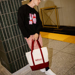 A person standing in a subway station holding a white canvas tote bag with a dark red bottom panel and handles. The person is wearing a black graphic sweatshirt, pink striped pants, white sneakers, and a purple cap.
