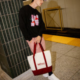 A person standing in a subway station holding a white canvas tote bag with a dark red bottom panel and handles. The person is wearing a black graphic sweatshirt, pink striped pants, white sneakers, and a purple cap.