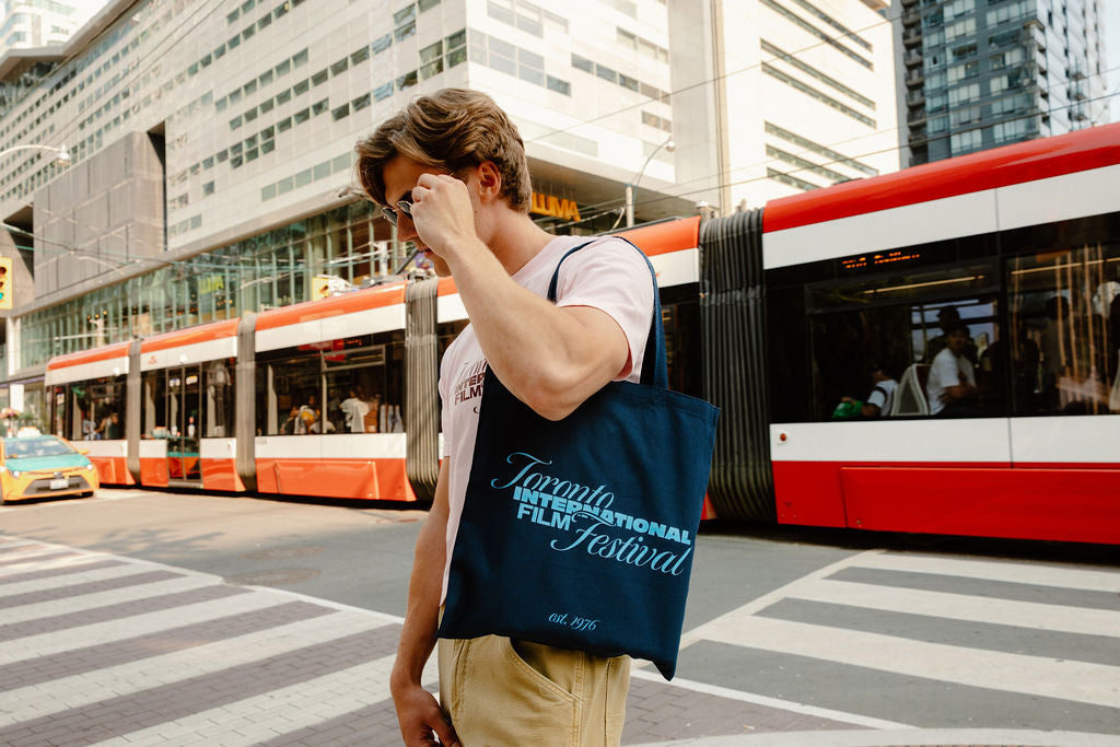 A man wearing sunglasses and a light pink t-shirt walks across a city street crosswalk, carrying a navy blue tote bag printed with the text 'Toronto International Film Festival est. 1976'.