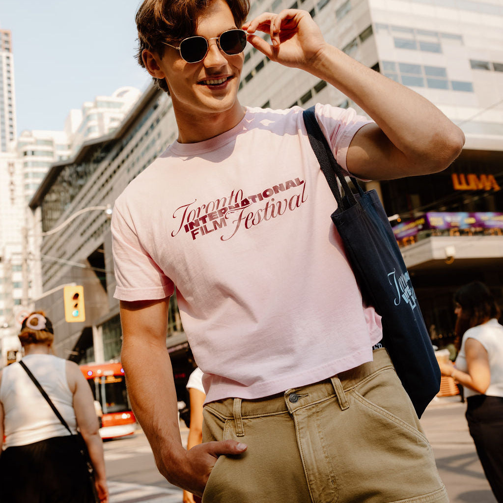 A man wearing a light pink graphic t-shirt with 'Toronto International Film Festival' printed on the chest, khaki cargo pants, and sunglasses, standing on a city street with a blue tote bag.