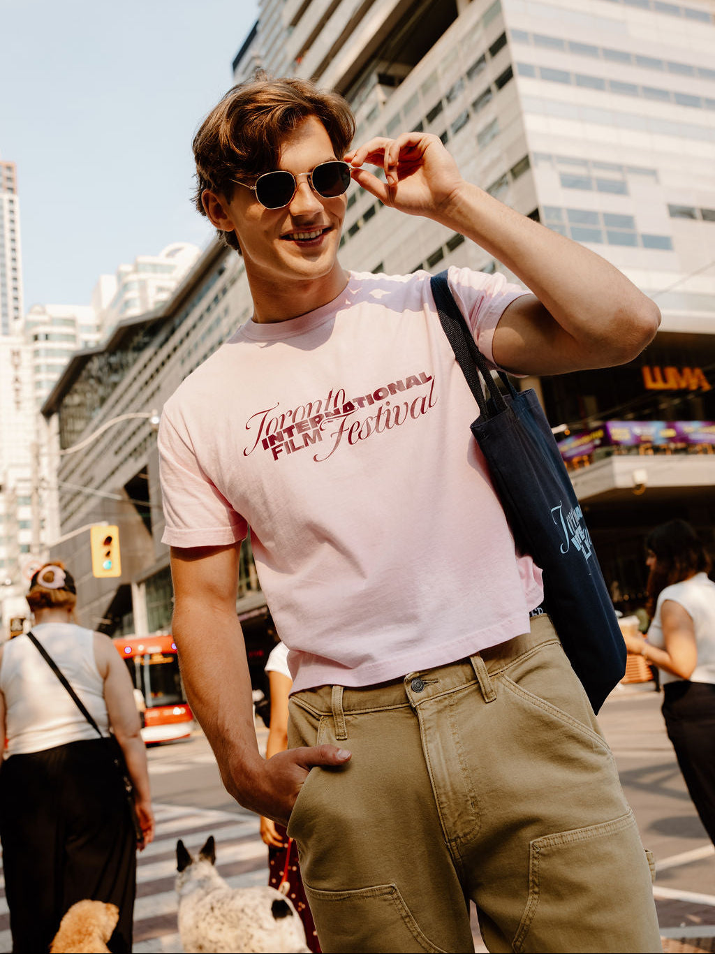 A man wearing a light pink graphic t-shirt with 'Toronto International Film Festival' printed on the chest, khaki cargo pants, and sunglasses, standing on a city street with a blue tote bag.