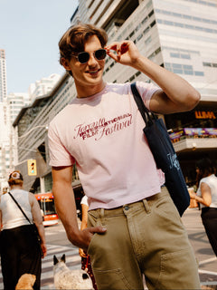 A man wearing a light pink graphic t-shirt with 'Toronto International Film Festival' printed on the chest, khaki cargo pants, and sunglasses, standing on a city street with a blue tote bag.