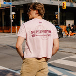 A man standing on a city street corner wearing a light pink graphic t-shirt with 'SEPTEMBER 10 20' printed in maroon on the back, paired with khaki cargo pants.