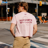 A man standing on a city street corner wearing a light pink graphic t-shirt with 'SEPTEMBER 10 20' printed in maroon on the back, paired with khaki cargo pants.