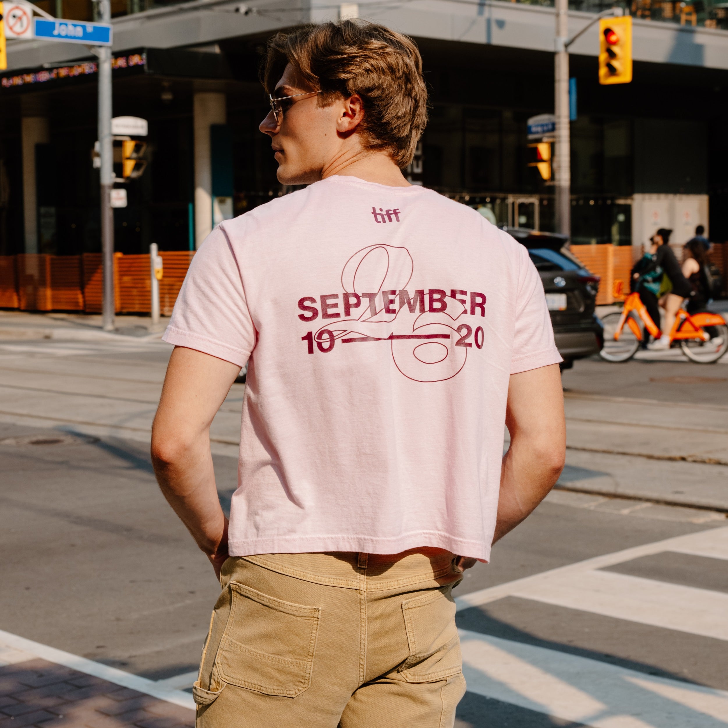 A man standing on a city street corner wearing a light pink graphic t-shirt with 'SEPTEMBER 10 20' printed in maroon on the back, paired with khaki cargo pants.